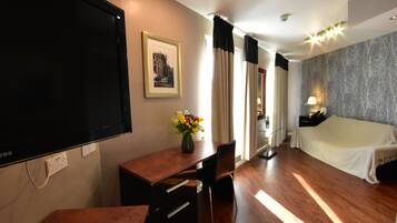 A hotel room with a bed, a desk with a chair, a TV, a framed picture, and a vase with flowers.