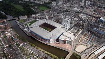 Vista aérea de un estadio con un campo central amplio, rodeado de una combinación de edificios residenciales y comerciales, y un río que corre paralelo al estadio.