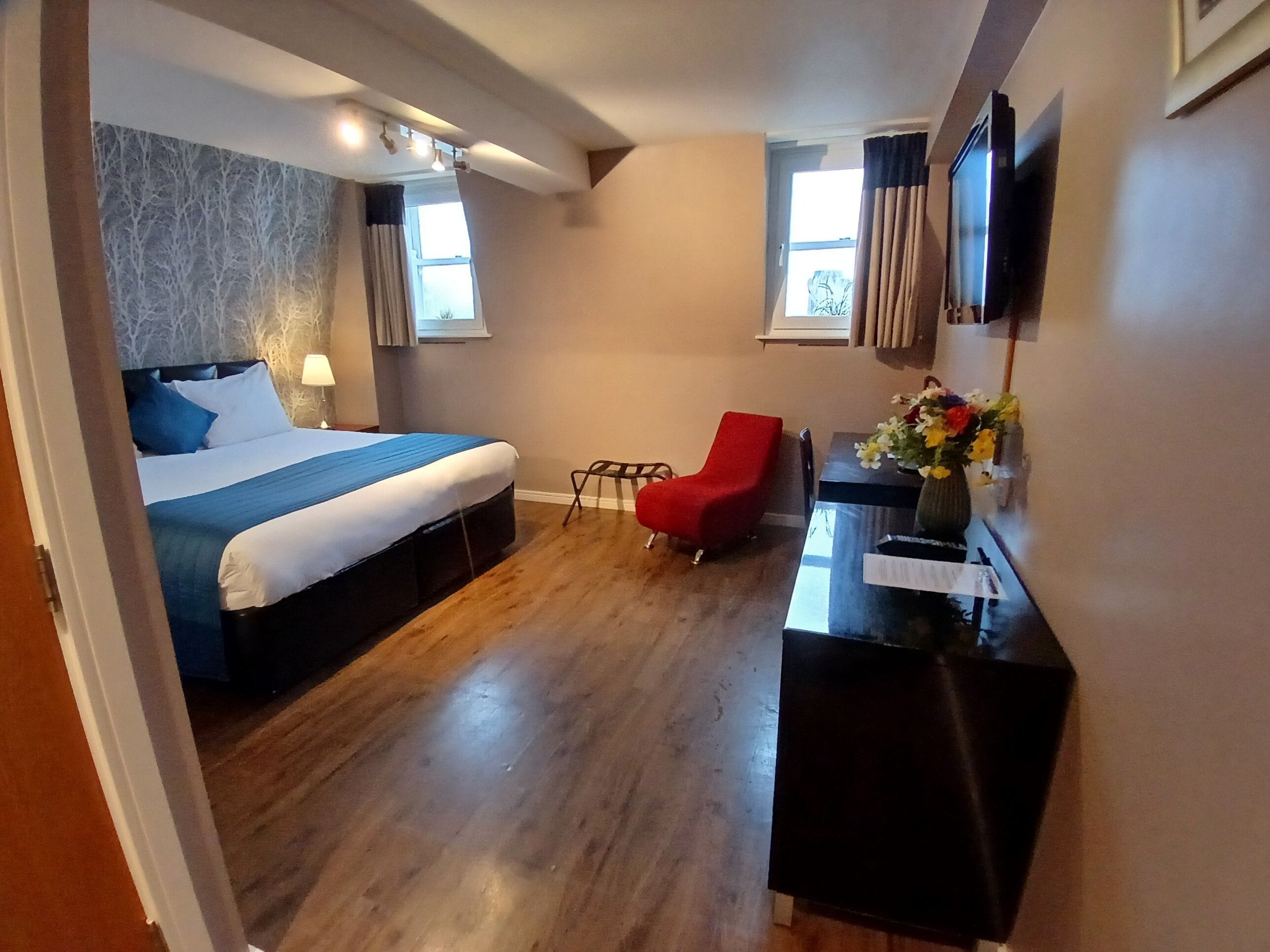 Quarto de hotel com cama, uma cadeira vermelha, escrivaninha com um vaso de flores e televisão na parede.