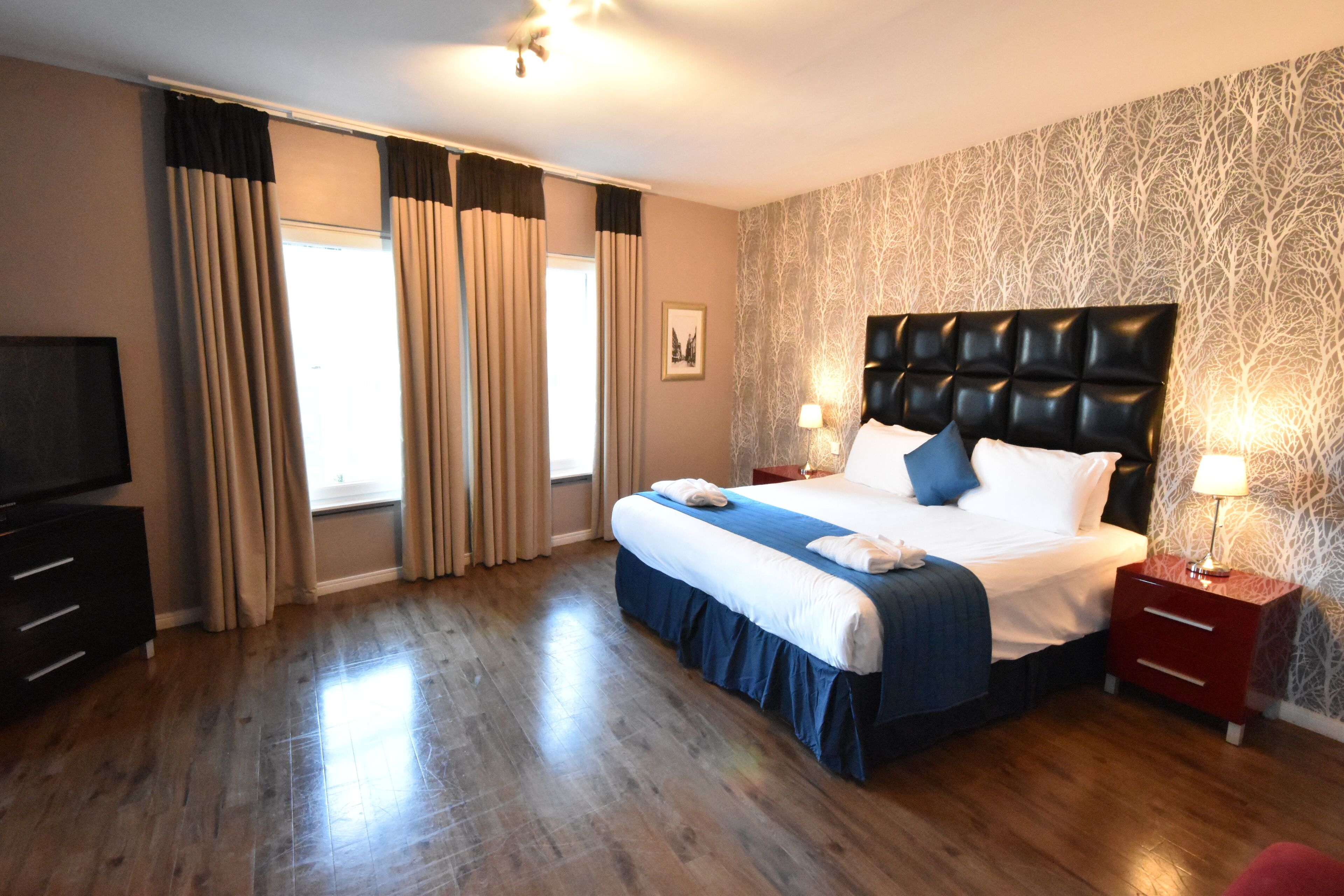 A hotel room with a large bed, a TV, a red nightstand, and a patterned wallpaper.