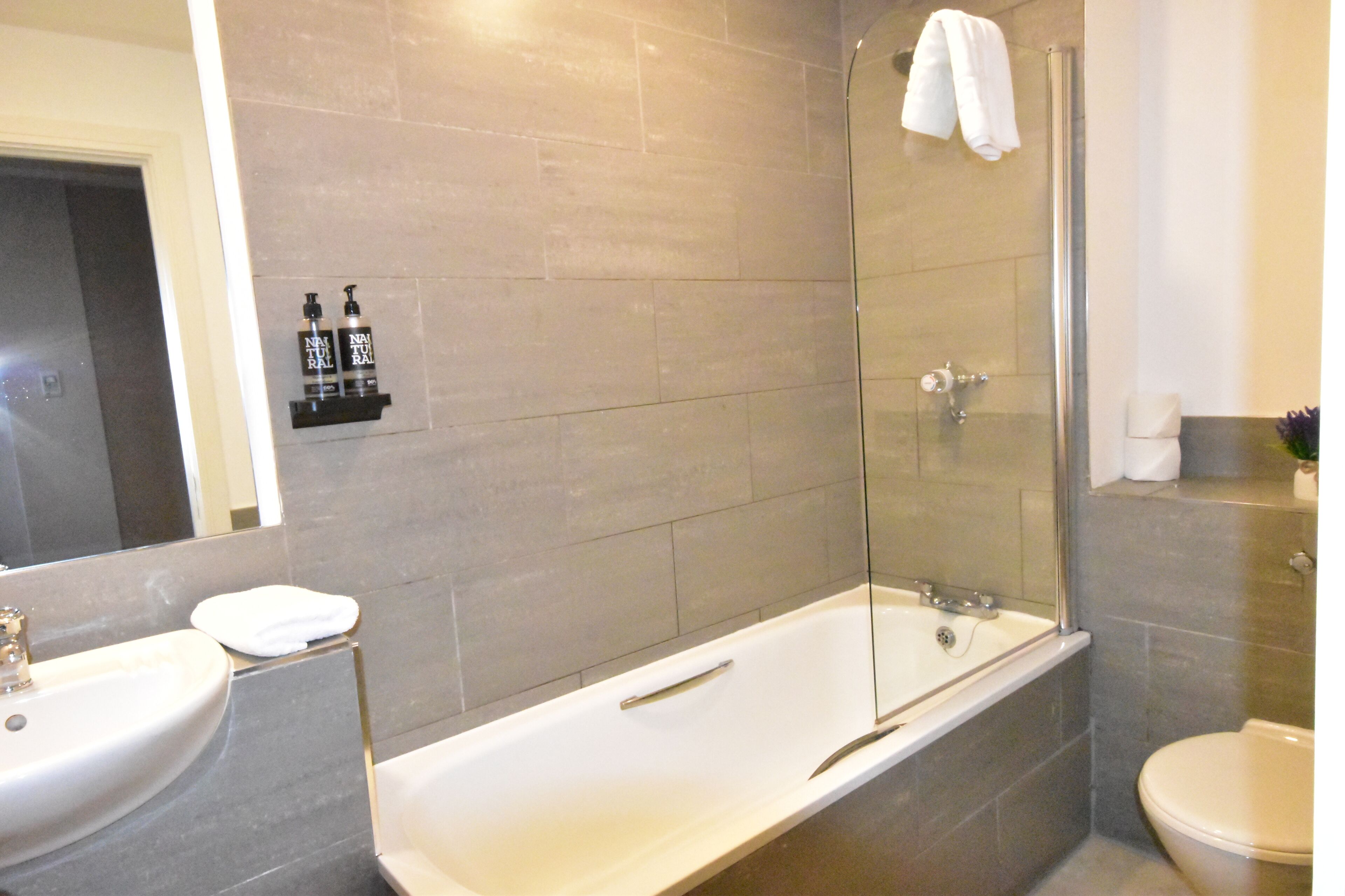 Een moderne badkamer met een bad, een wastafel en een toilet. Op een plank staan twee flessen shampoo.