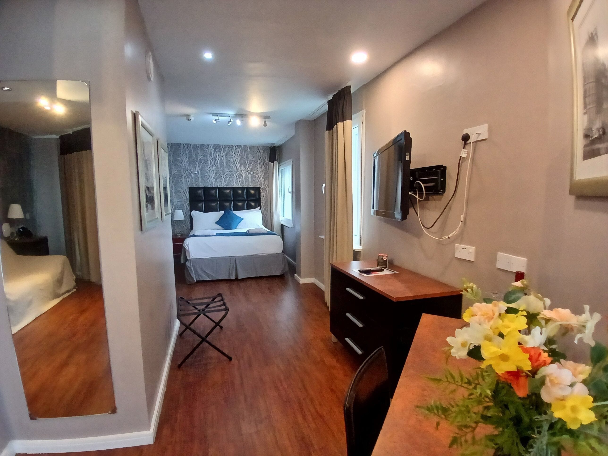 Ein Hotelzimmer mit Bett, Schreibtisch, Stuhl, Spiegel und einer Blumenvase.