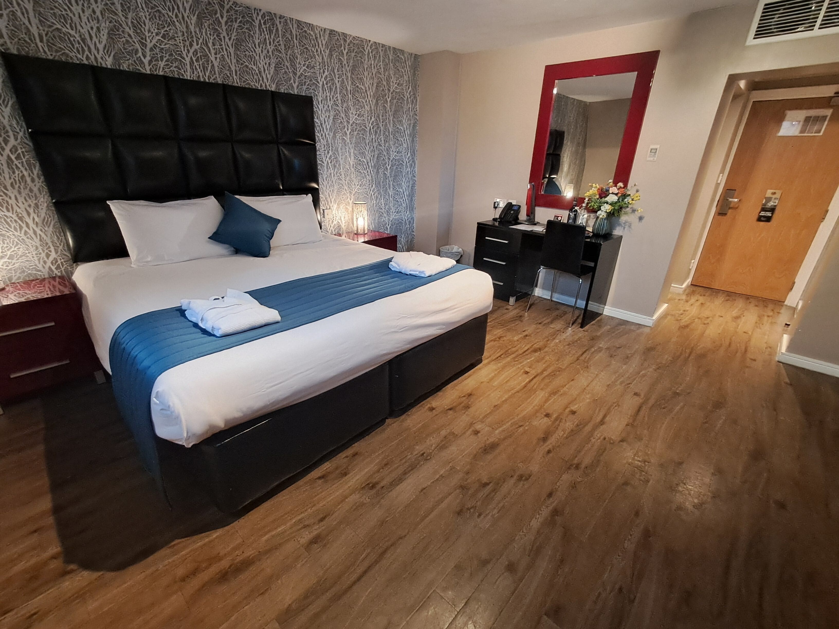 A neatly arranged hotel room with a large bed, a desk, a mirror, and a wooden floor.