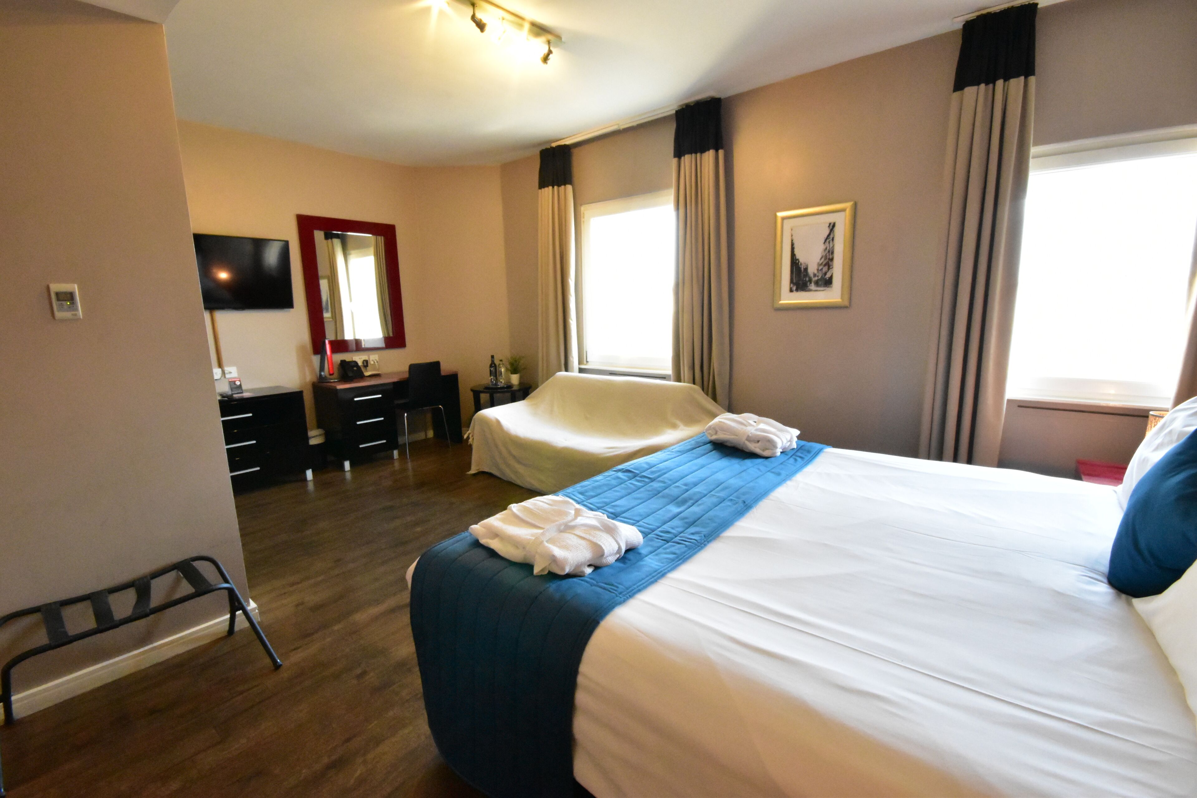 Una habitación de hotel con cama, escritorio, silla, televisor, ventana con cortinas y una mesita con una botella y una planta.