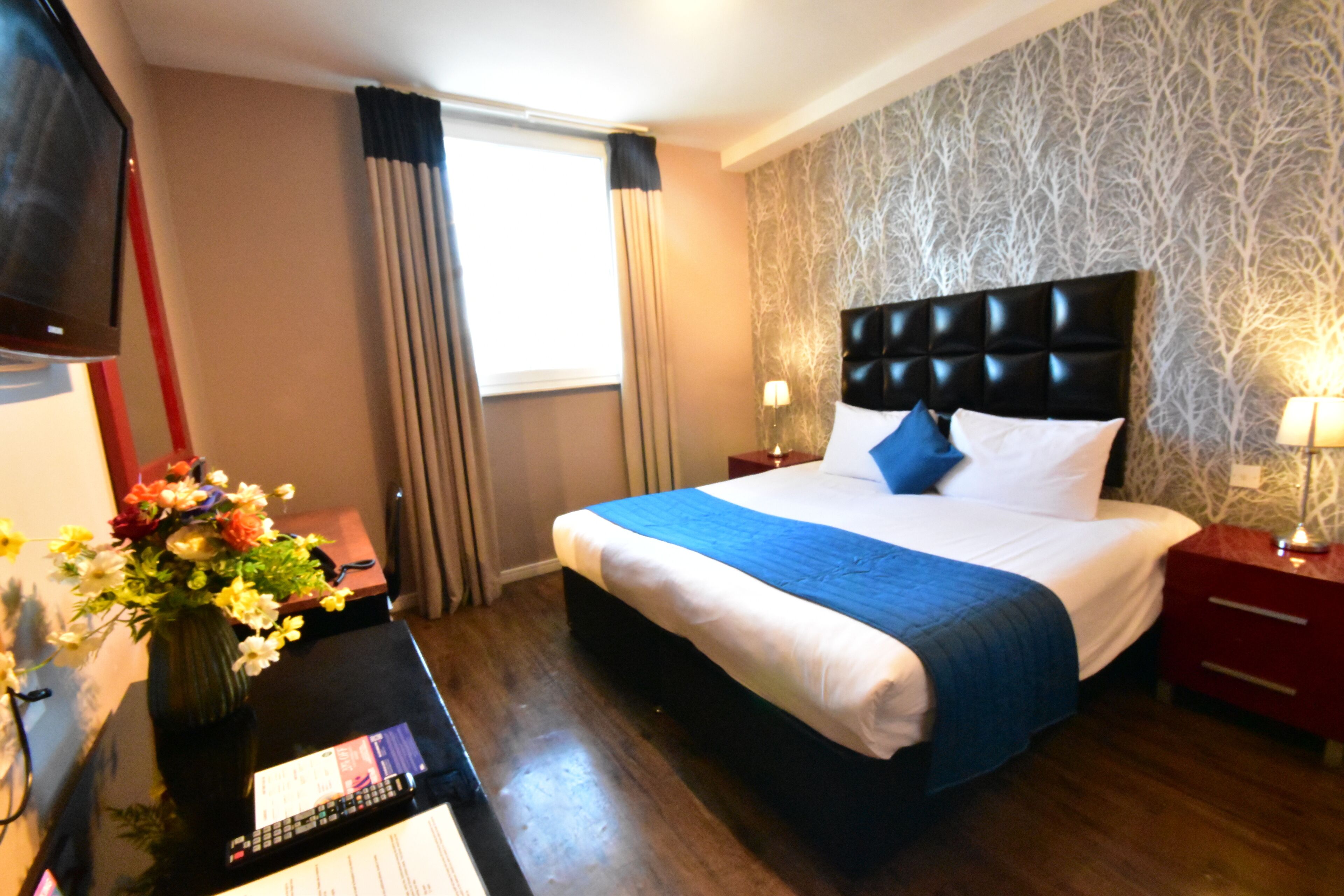 A hotel room with a large bed, a TV, a vase with flowers, and a desk with a lamp.