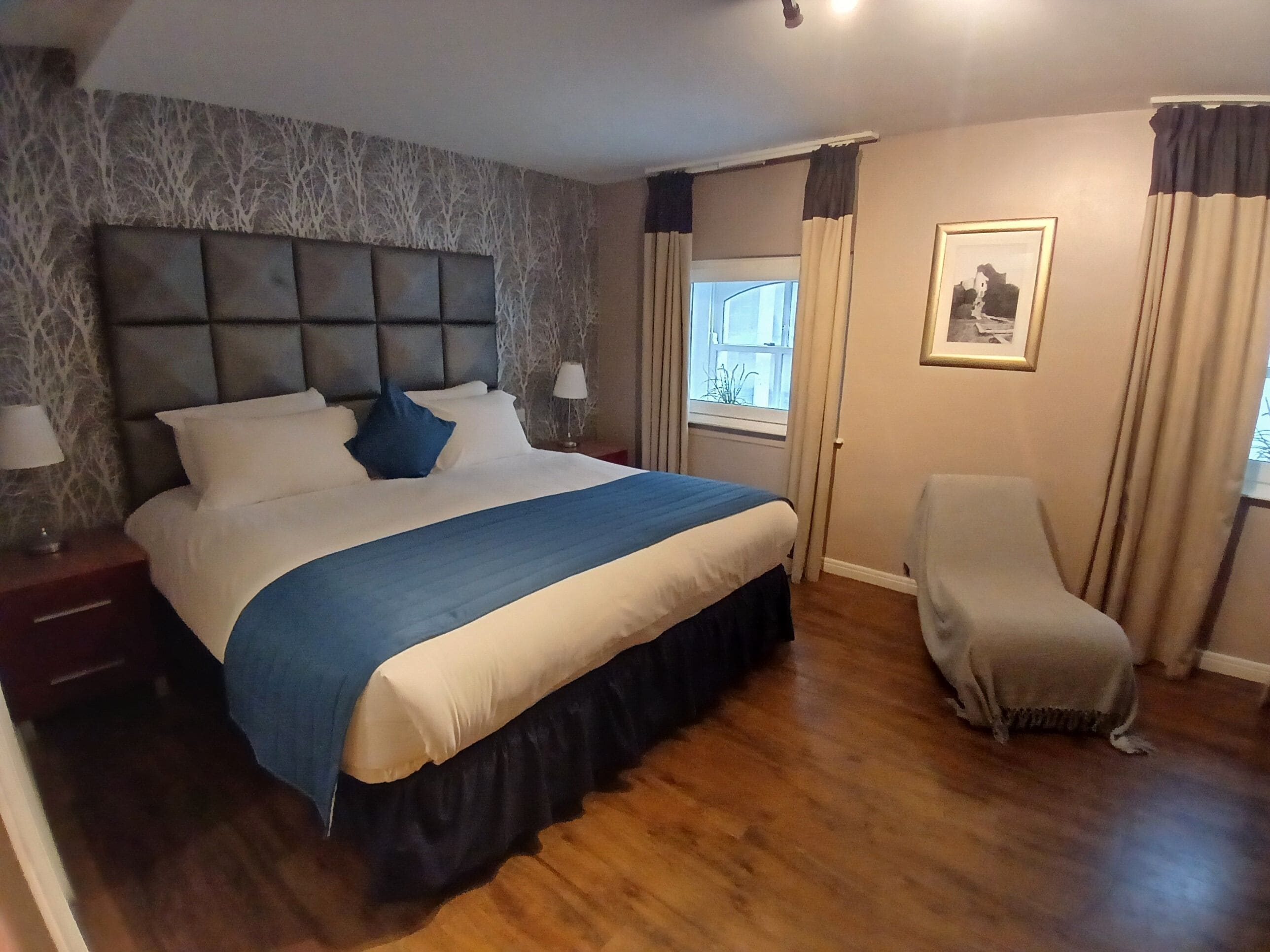 Un dormitorio con una cama grande, una mesita de noche, una silla, una ventana con cortinas y un cuadro en la pared.