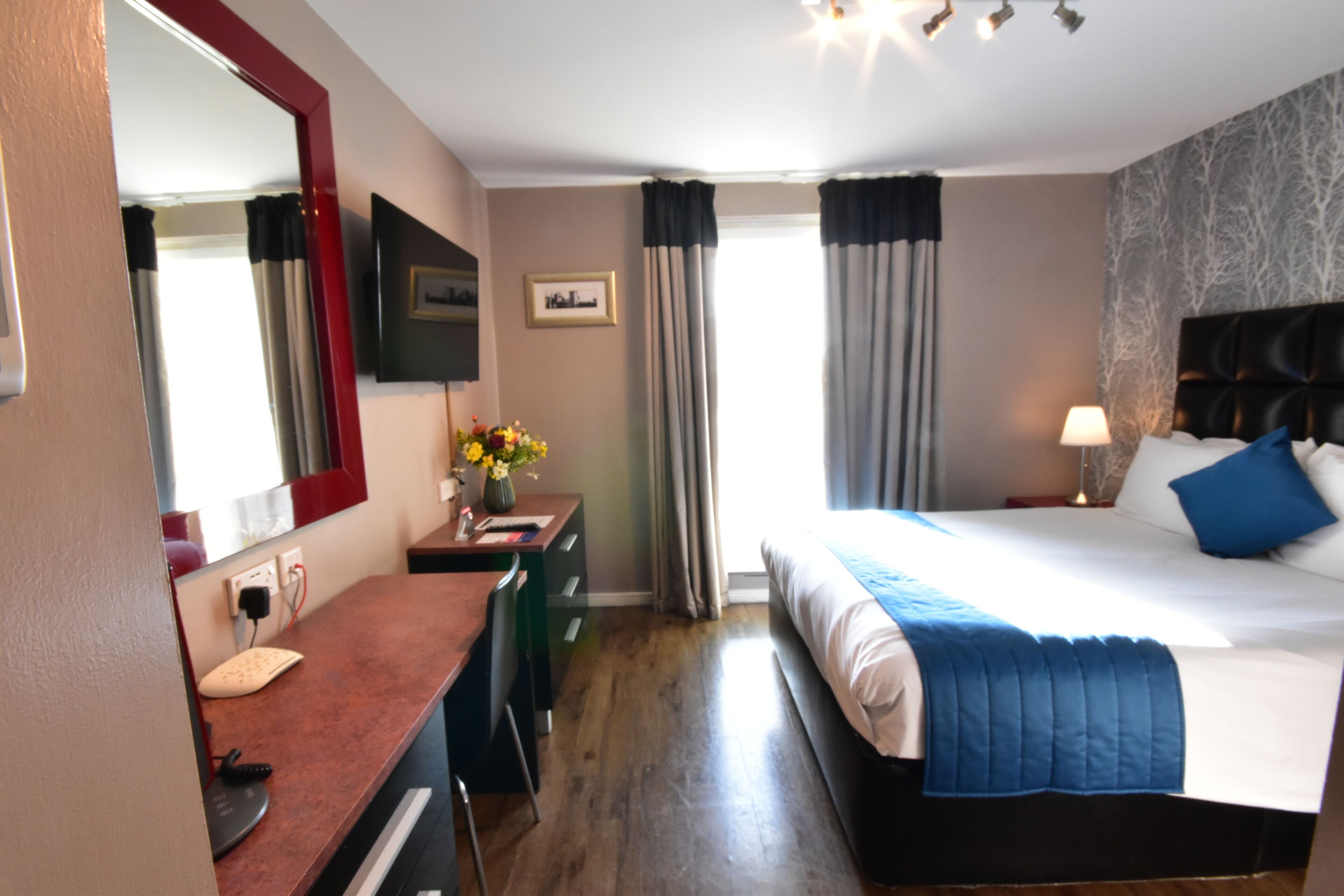 Quarto de hotel com cama, escrivaninha, cadeira, espelho, janela com cortinas e televisão fixada na parede.