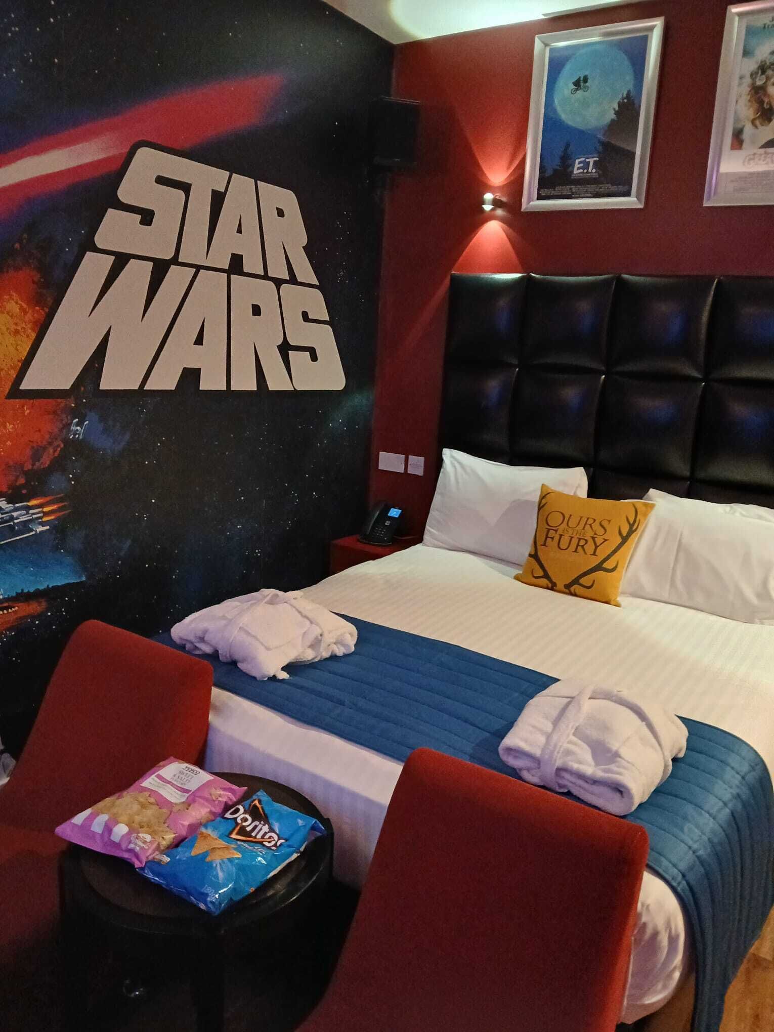 A Star Wars-themed hotel room with a large wall mural, a tufted headboard, a bed with white and blue bedding, a pillow with a quote, a small table with snacks, and a chair.