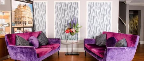 A reception area with two purple velvet sofas, a glass coffee table, and a decorative floral arrangement.