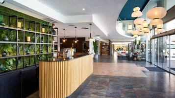 Um saguão de hotel com balcão de recepção, uma parede verde com plantas e uma área de estar com mesas e cadeiras.