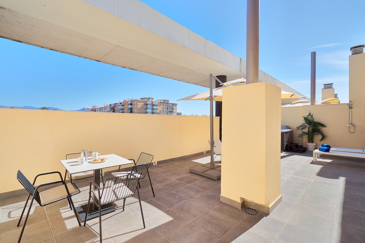 Una terraza en la azotea con una mesa, sillas, una planta en maceta y vistas a edificios al fondo.
