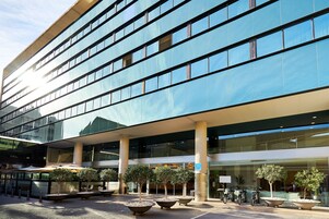 Un edificio moderno con fachada de vidrio, zona de estar al aire libre con sombrillas y espacio para aparcar bicicletas.