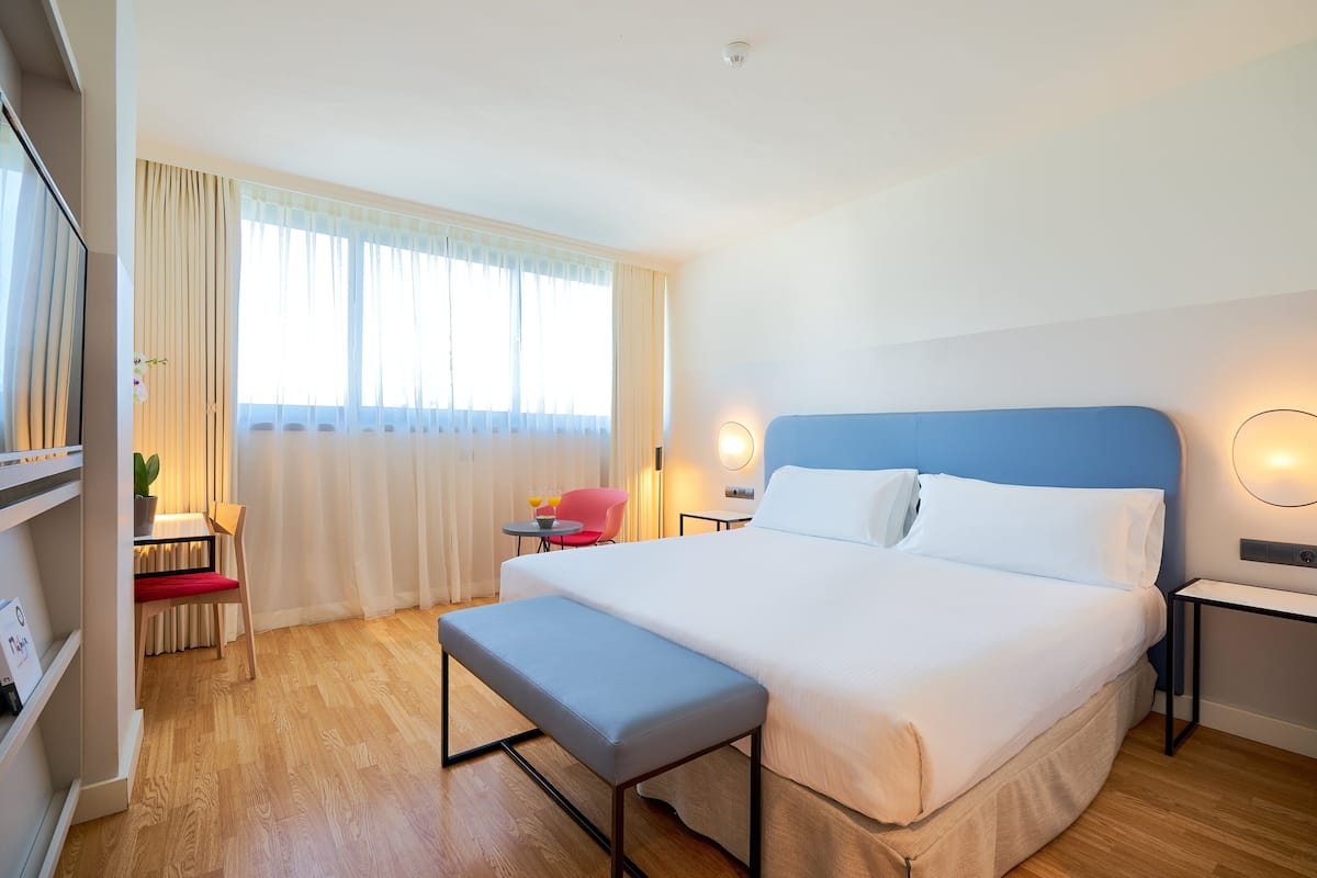 Habitación de hotel con una cama grande, un banco, un escritorio con una silla, una mesita con una planta y una ventana con cortinas.