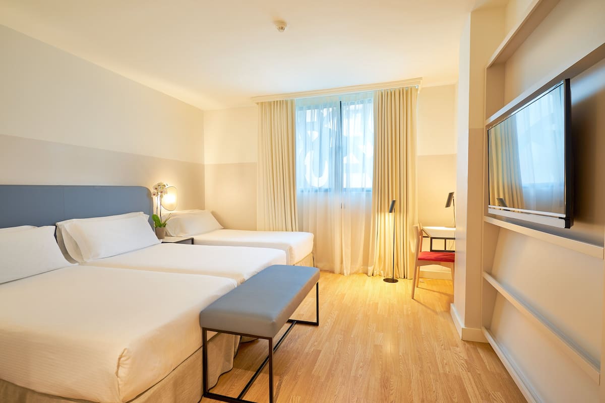 Habitación de hotel con dos camas, un banco, un escritorio, un televisor y una ventana con cortinas.