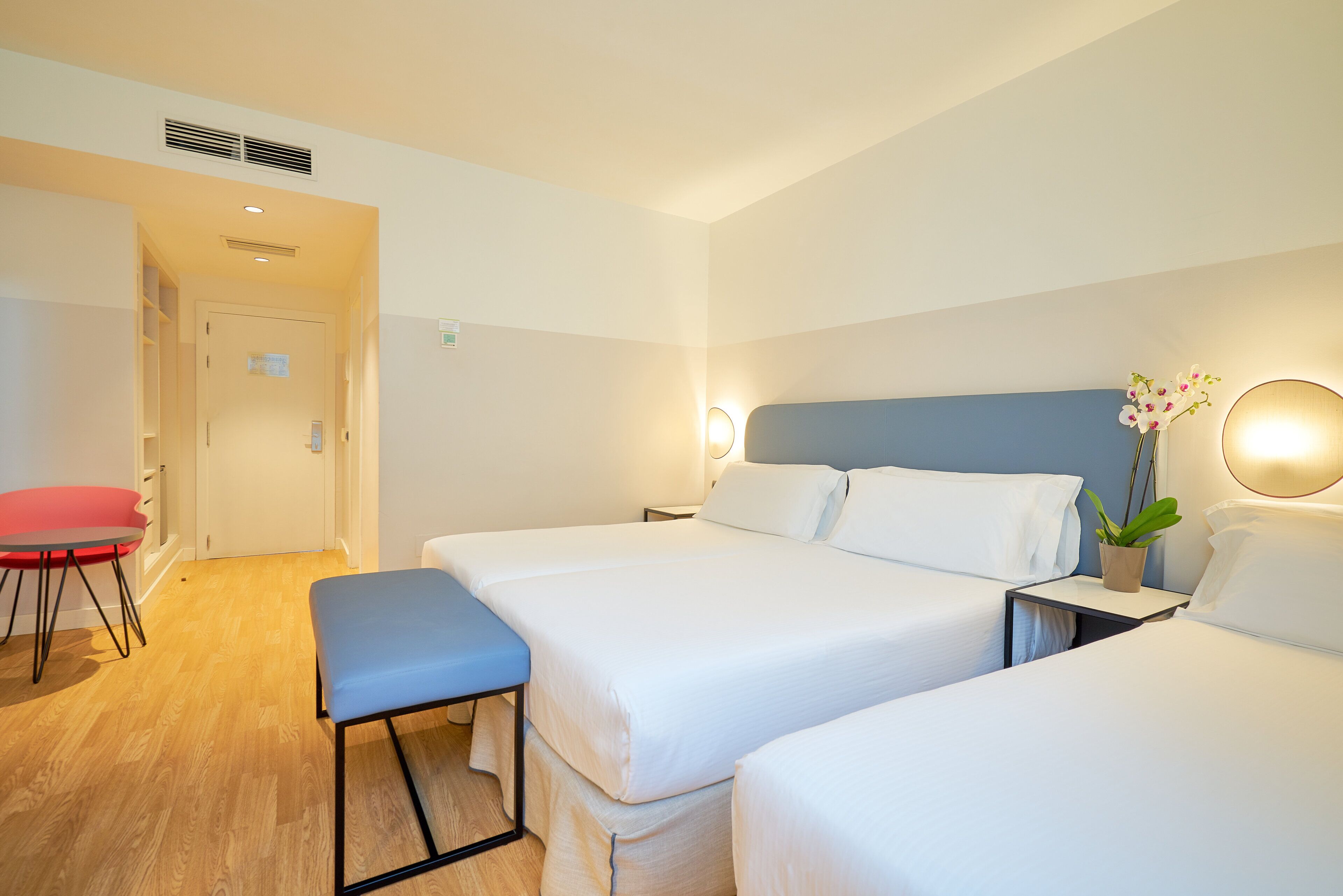 A hotel room with two beds, a desk, a chair, and a small table with a plant.