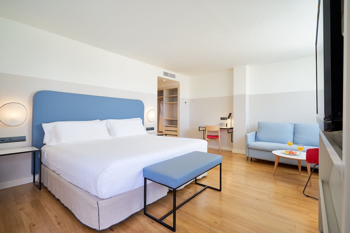 Una habitación de hotel moderna con una cama grande, un banco, un sofá, una mesita con un plato y vasos, y un armario.