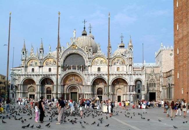 A historic building with multiple arches and a central dome, surrounded by a plaza with pigeons.