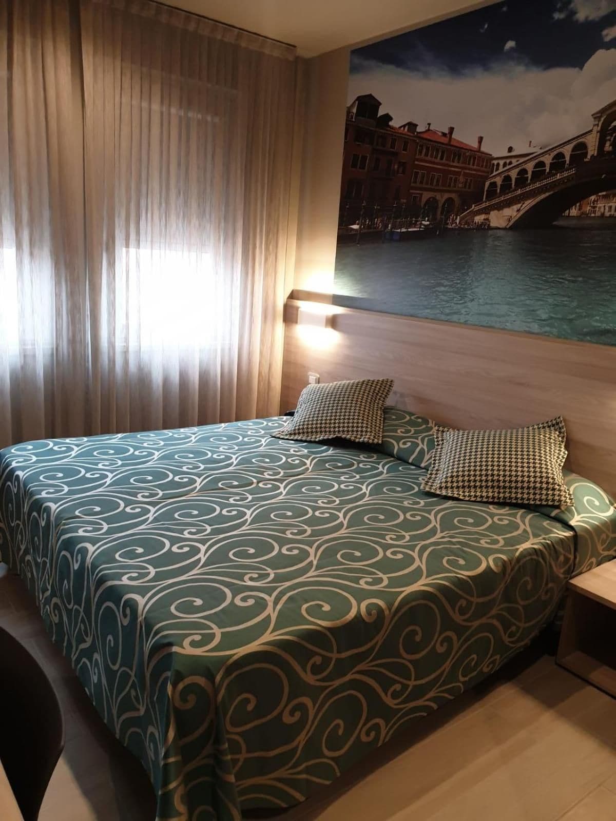 Habitación de hotel con cama, cortinas, luz nocturna y un mural de un paisaje urbano.