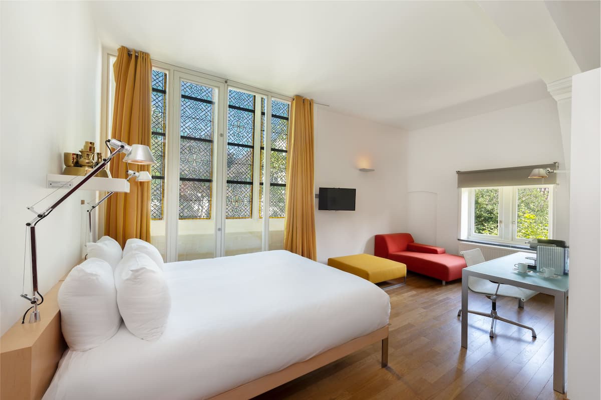 Habitación de hotel con una cama grande, un escritorio con silla, un sofá rojo y un televisor en la pared.