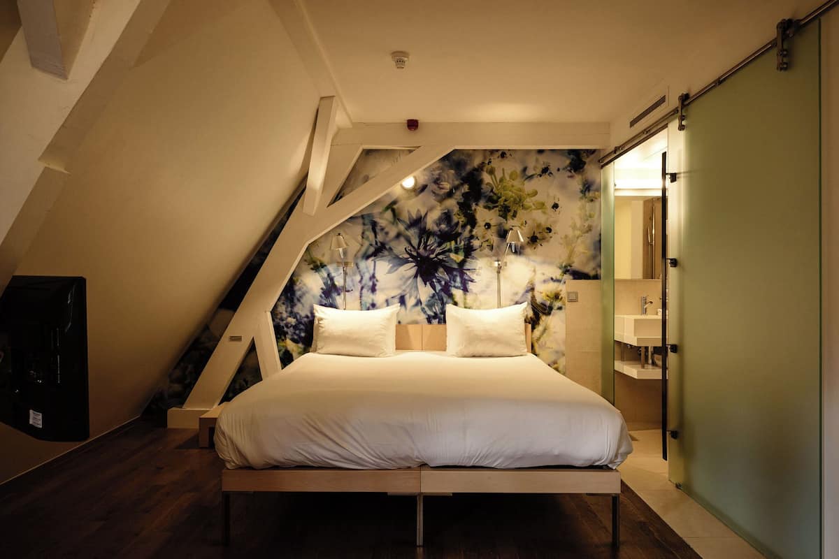 Un dormitorio moderno con techo inclinado, una cama grande con ropa de cama blanca y un diseño de papel tapiz floral.