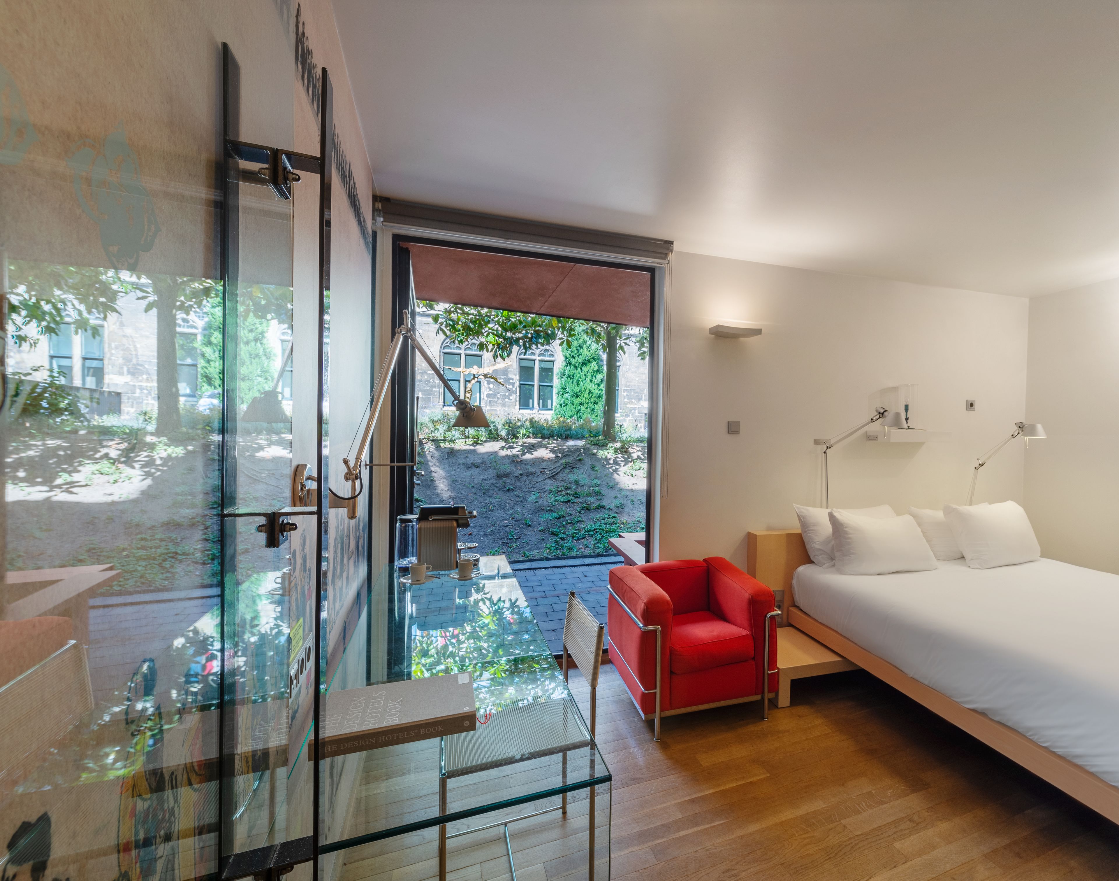 A modern hotel room with a glass door leading to an outdoor area, a red chair, a bed with white linens, and a desk with a lamp.