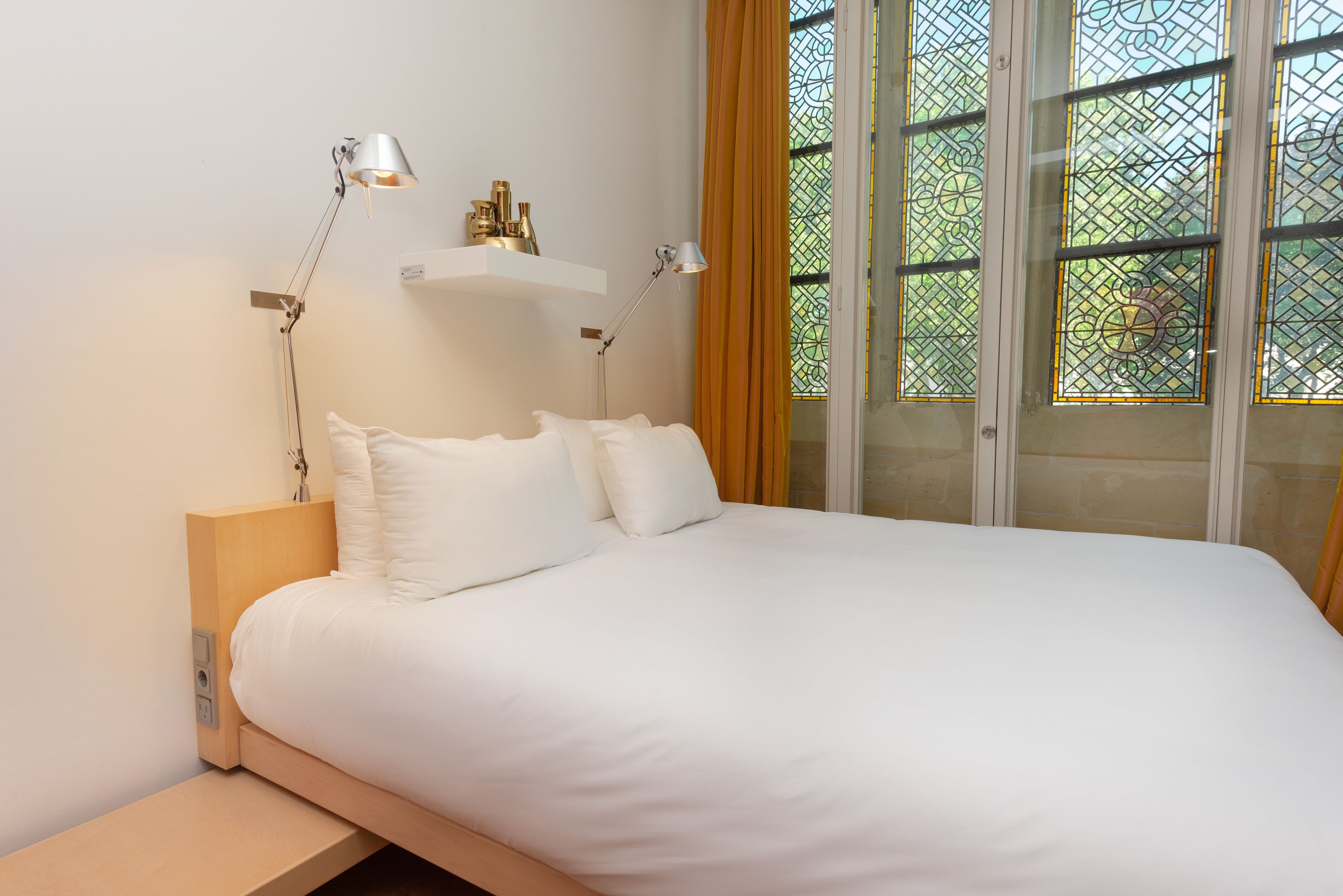 Een net opgemaakt bed met witte lakens en twee bedlampjes. Daarboven bevinden zich twee wandplanken, en een raam met uitzicht op groen.