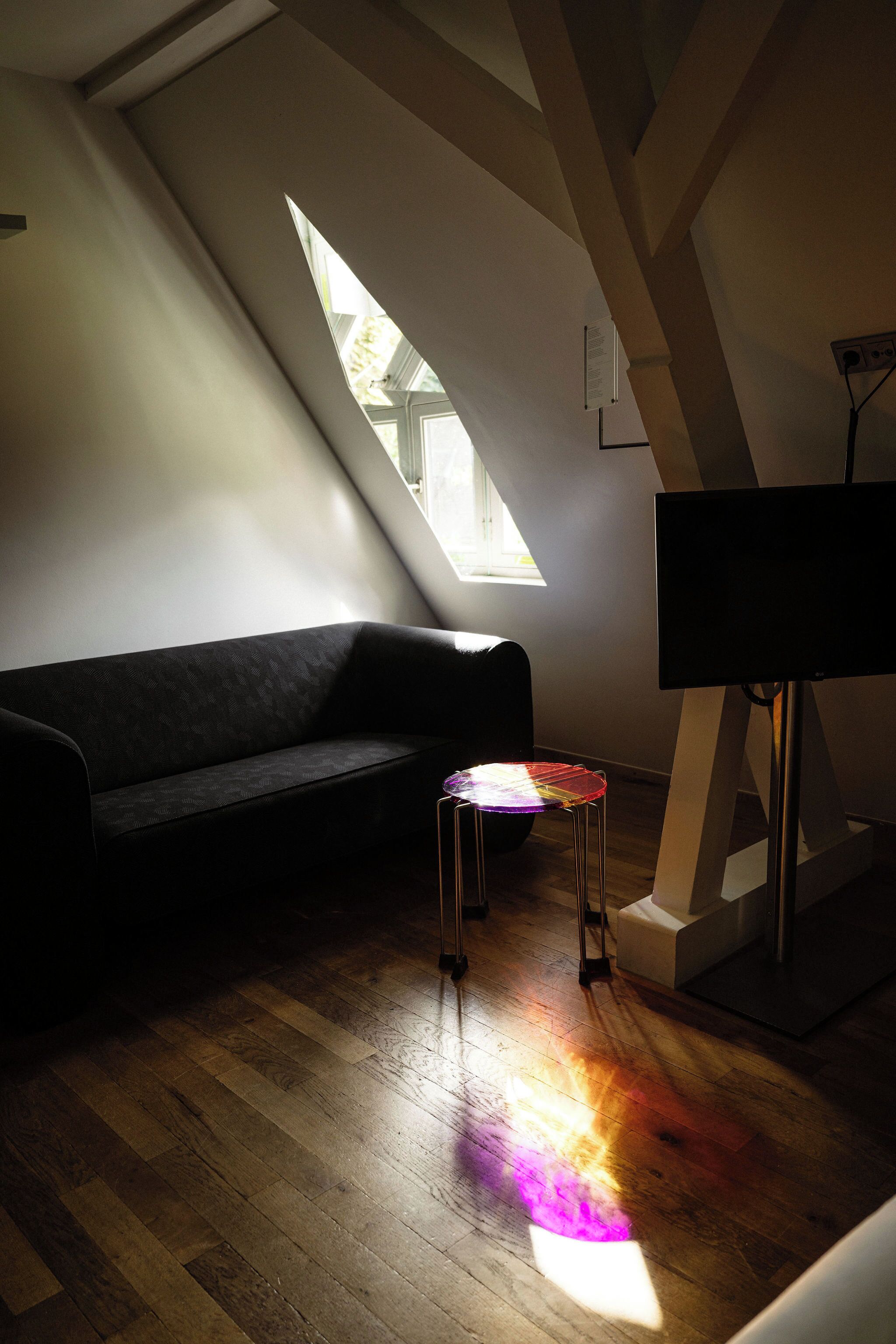 Ett rum med ett sluttande tak, en soffa i mörk färg, en liten pall och en platt-TV som står på ett stativ.