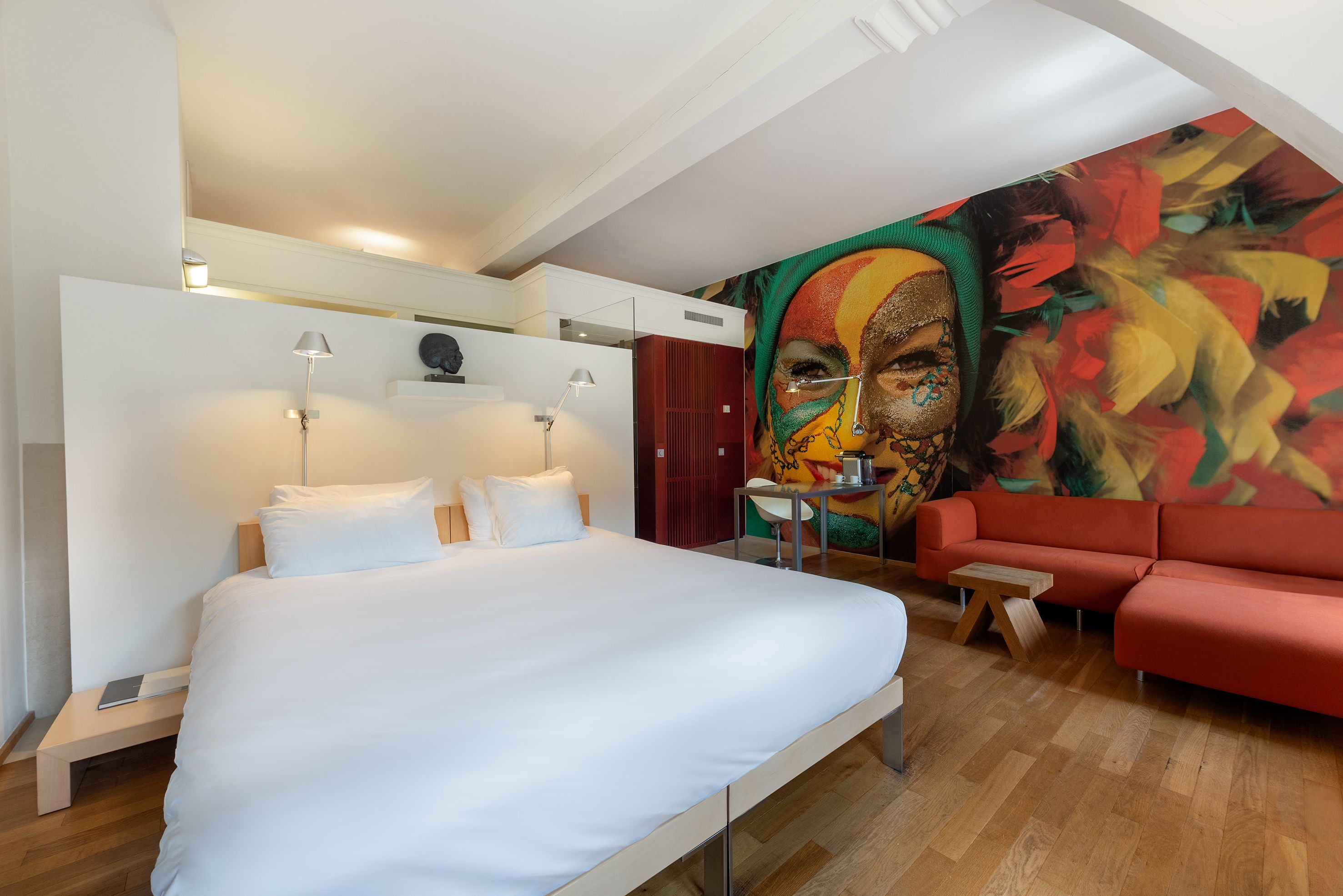 Un dormitorio moderno con una cama grande, un sofá rojo, una mesita y un mural de colores en la pared.