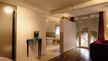A modern hotel room with a wooden floor, a red chair, a white sofa, a glass partition, and a bathroom with a bathtub and a sink.