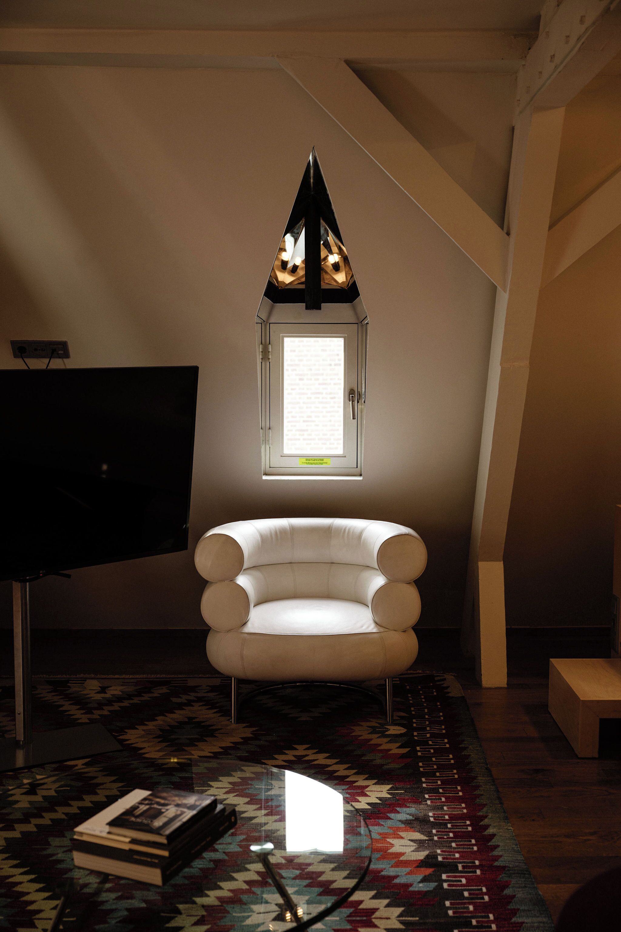 A room with a white armchair, a flat-screen TV, a patterned rug, and a window with a view of the outside.