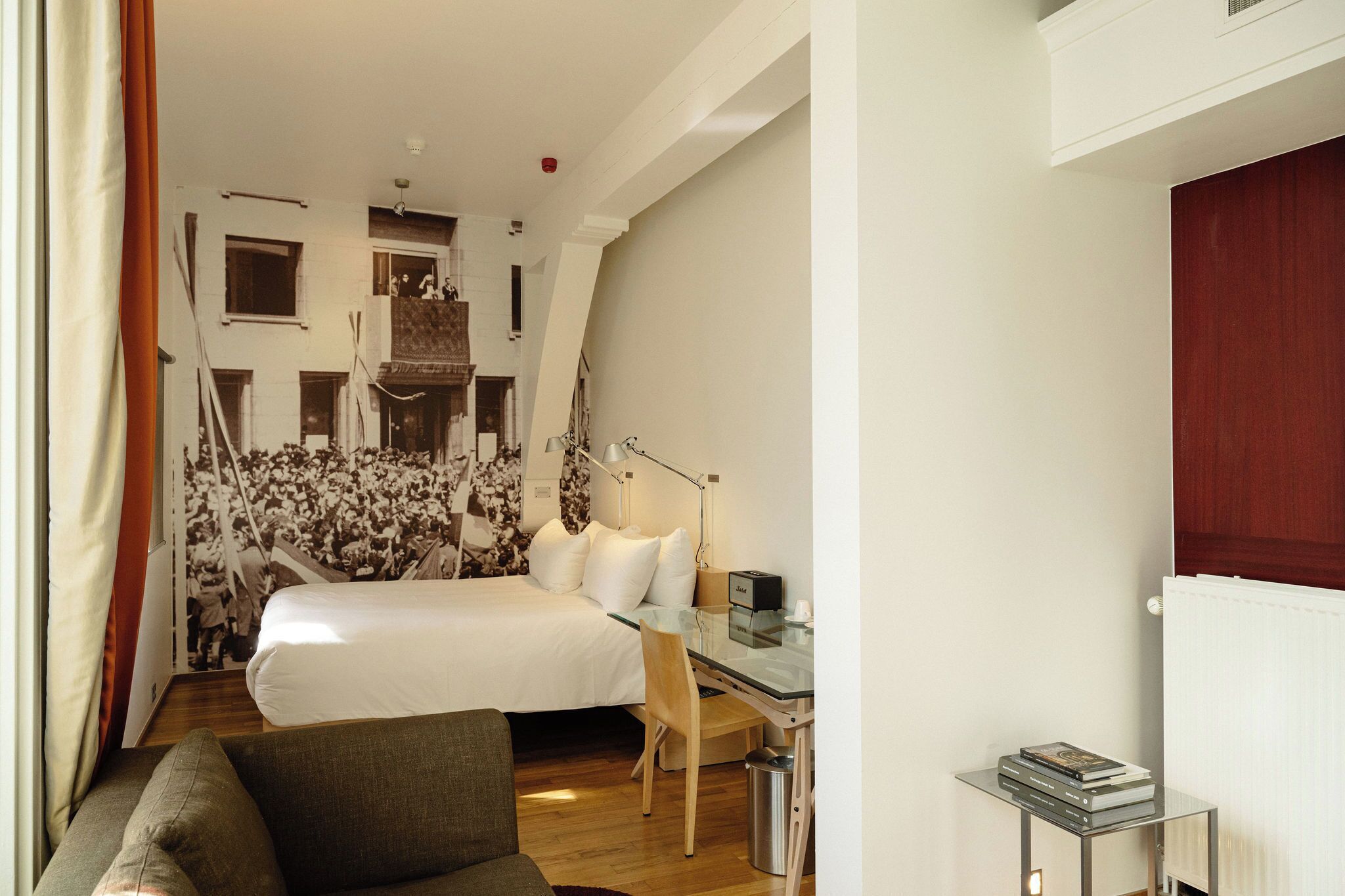 A modern hotel room with a large bed, a glass desk, and a view of a cityscape through the window.