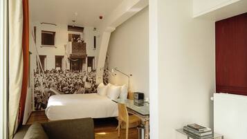 A modern hotel room with a large bed, a glass desk, and a view of a cityscape through the window.
