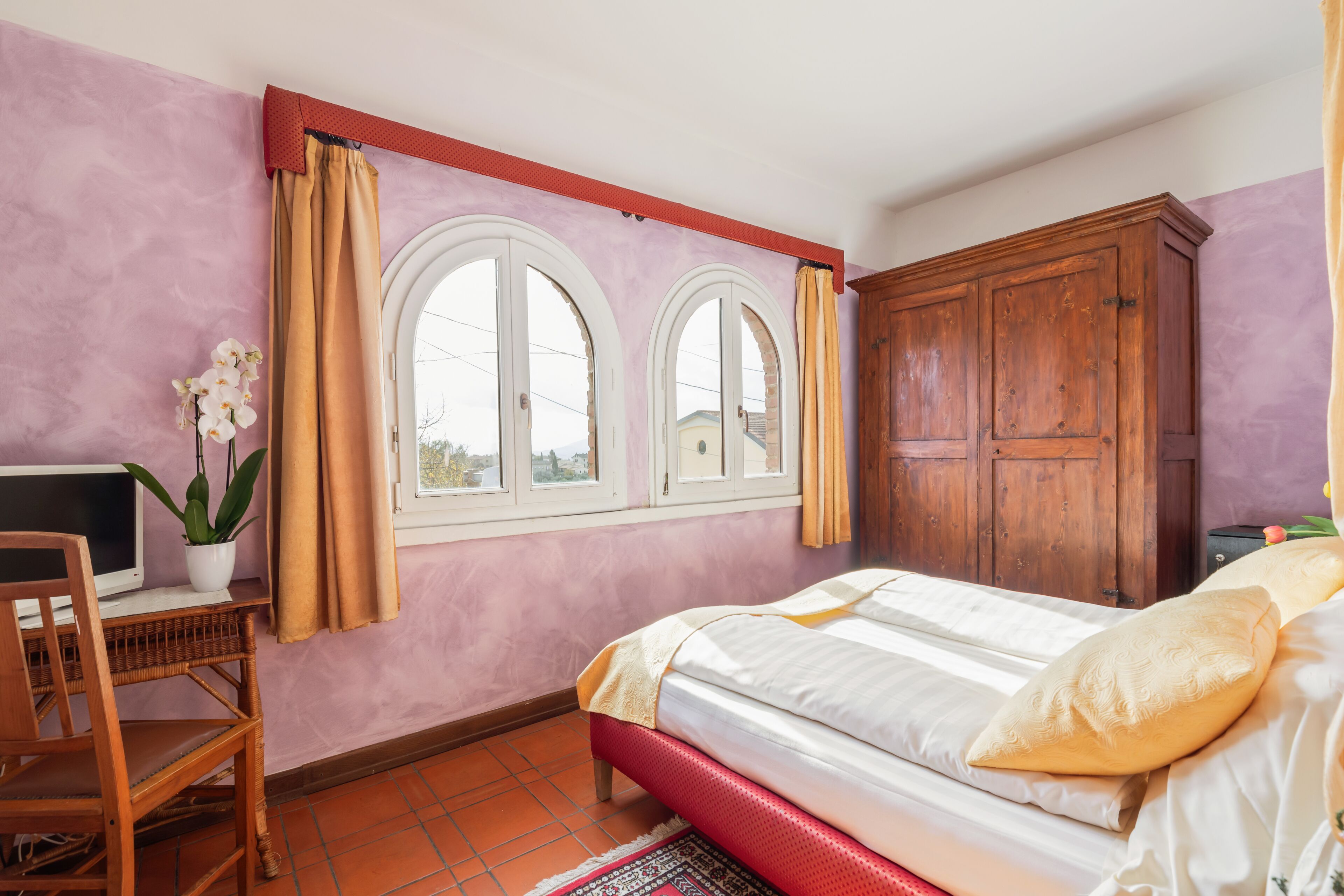 Ein Schlafzimmer mit einem Bett, einem Holzschrank, einem Schreibtisch mit Computer und einem Fenster mit Vorhängen.