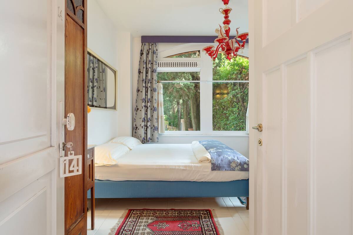 A bedroom with a bed, a window with curtains, a wooden door, and a red hanging light fixture.