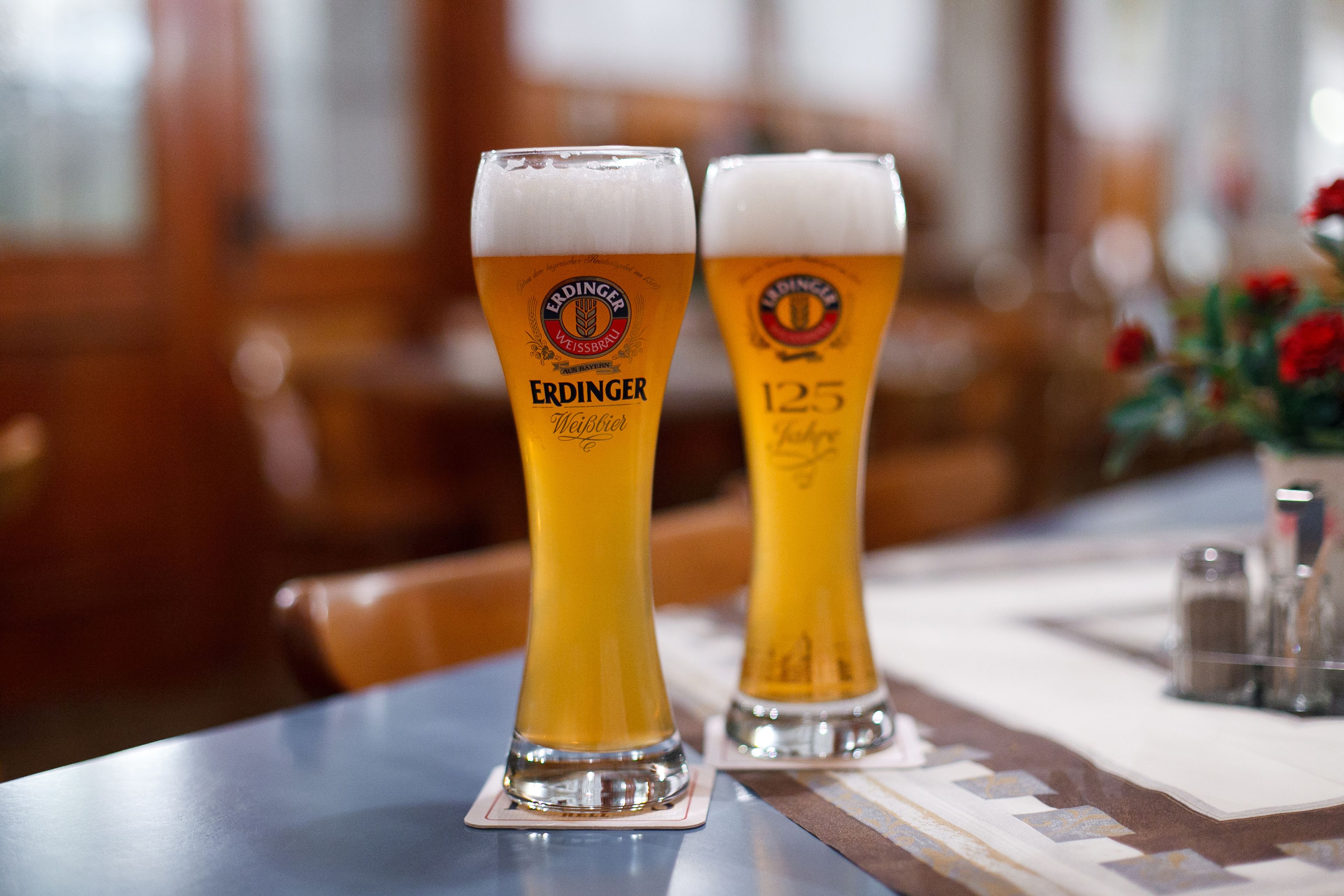 Two tall glasses of beer with the Erdinger Weihen logo on a table in a restaurant.