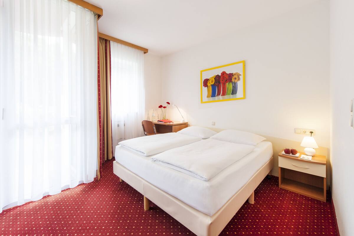 A hotel room with a double bed, a desk with a chair, a nightstand with a lamp, and a painting on the wall.