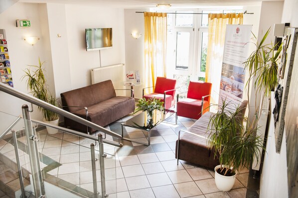 A hotel lobby with a brown sofa, red chairs, a glass coffee table with flowers, a potted plant, and a staircase with metal railings. - Andante Hotel Erding (Erding)