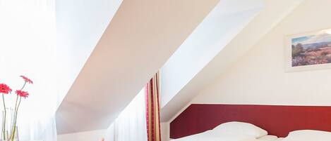 A bedroom with a red headboard, a bed with white linens, a window with sheer curtains, and a framed landscape painting on the wall.