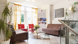 A hotel lobby with a reception desk, red and brown seating, potted plants, and a poster advertising European destinations.