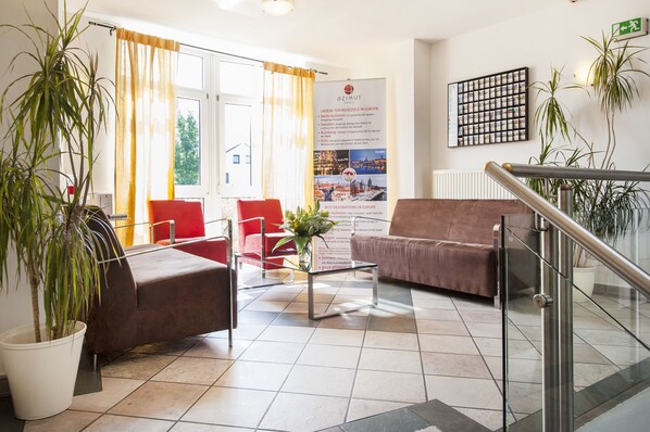 A hotel lobby with a reception desk, red and brown seating, potted plants, and a poster advertising European destinations. - Andante Hotel Erding (Erding)