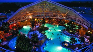 A large indoor swimming pool complex with multiple pools and lounge areas, surrounded by a glass-roofed structure.