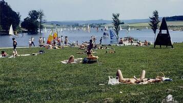 Eine bewachsene Wiese mit sonnenbadenden Personen und ein See mit Segelbooten im Hintergrund.