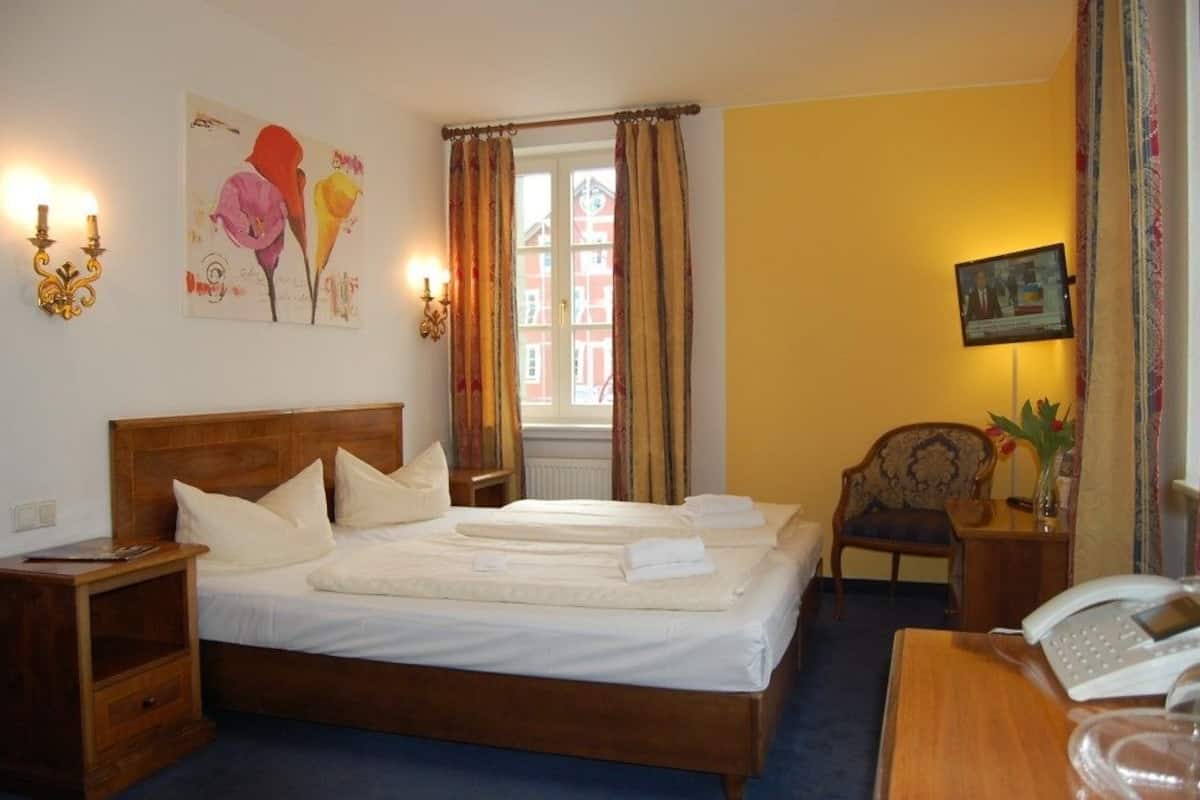 A hotel room with a wooden bed, a bedside table with a lamp, a chair, and a desk with a telephone.