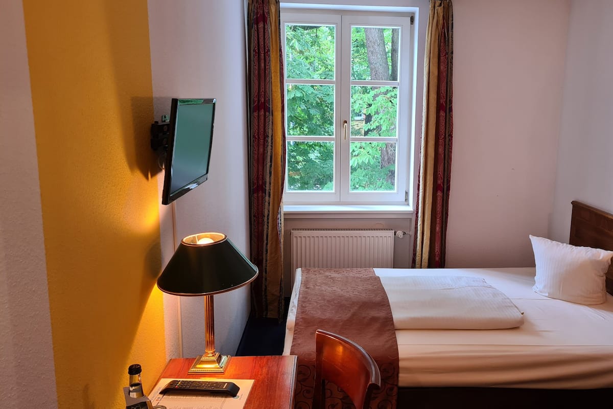 A hotel room with a bed, a desk with a lamp, a television mounted on the wall, and a window with curtains.