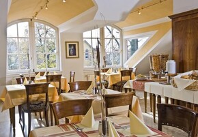 Un comedor con varias mesas y sillas, ventanas amplias con vista a árboles y un techo alto con vigas a la vista.