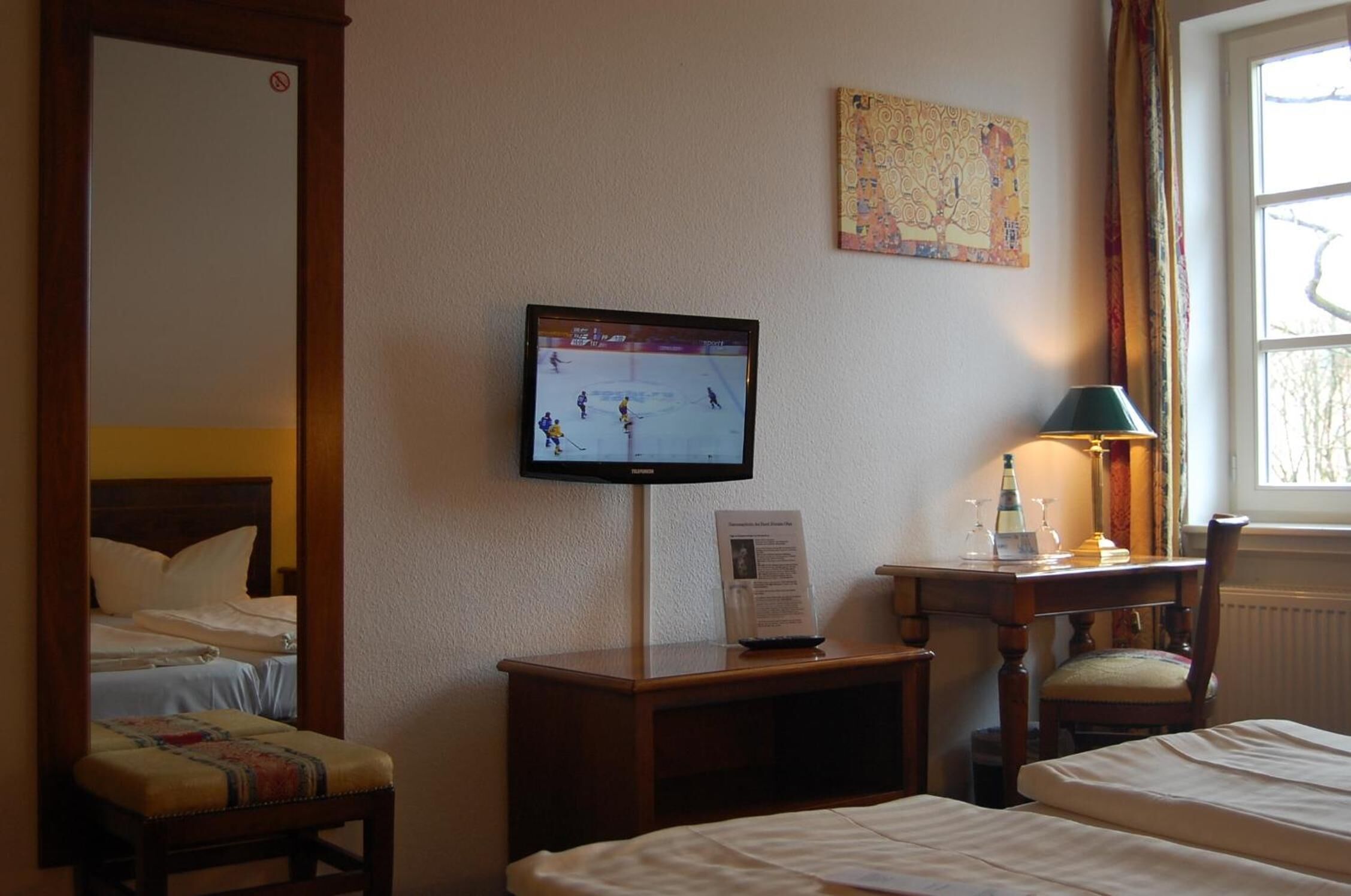 Ein Hotelzimmer mit Bett, Schreibtisch mit Lampe, Fernseher, der ein Eishockeyspiel zeigt, und einem Vorhangfenster.