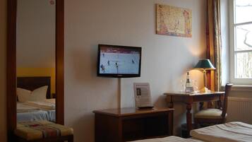 Ein Hotelzimmer mit Bett, Schreibtisch mit Lampe, Fernseher, der ein Eishockeyspiel zeigt, und einem Vorhangfenster.