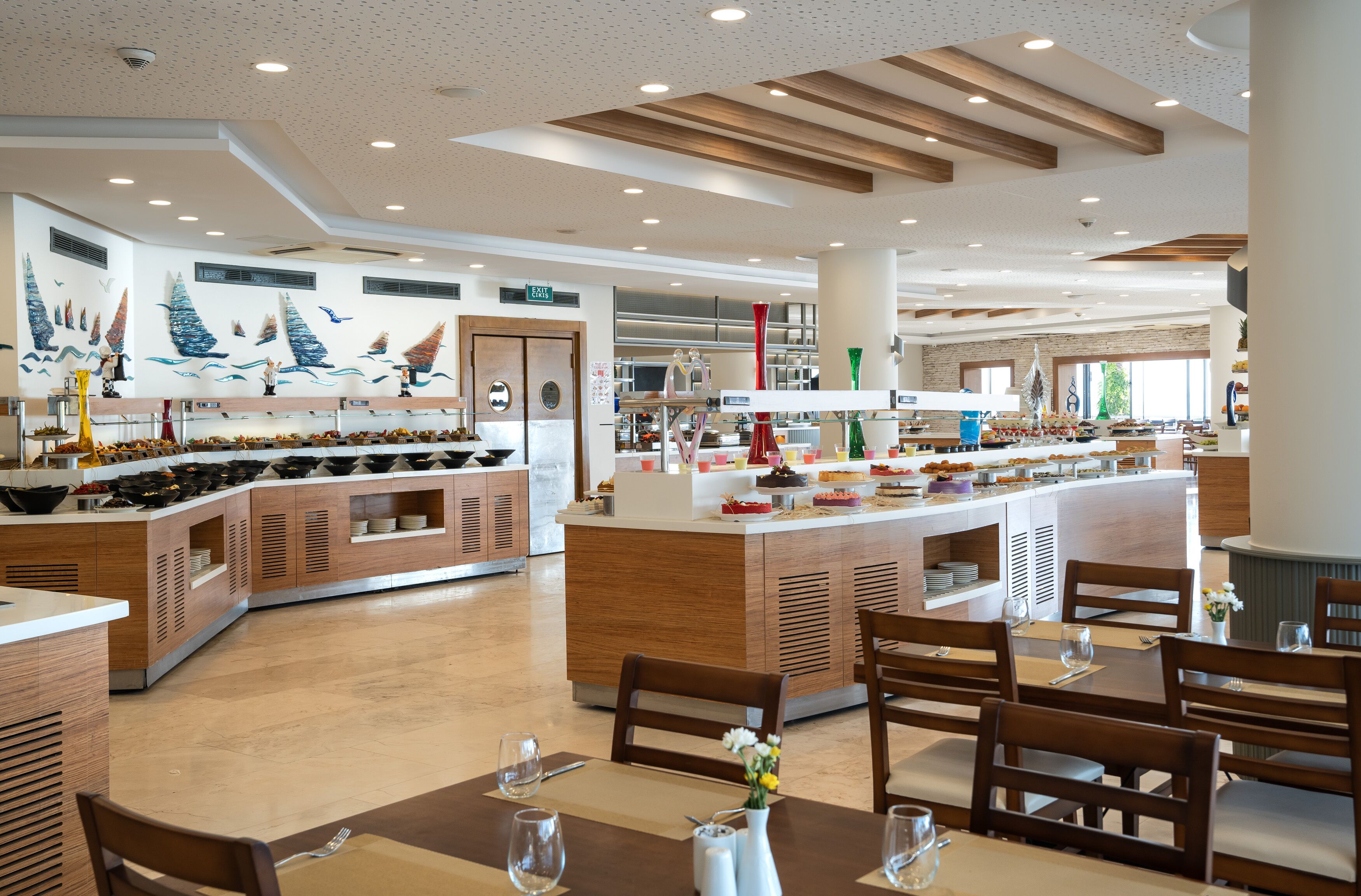 Un área de comedor de hotel moderna con una isla central, muebles de madera y un mural de veleros en la pared.