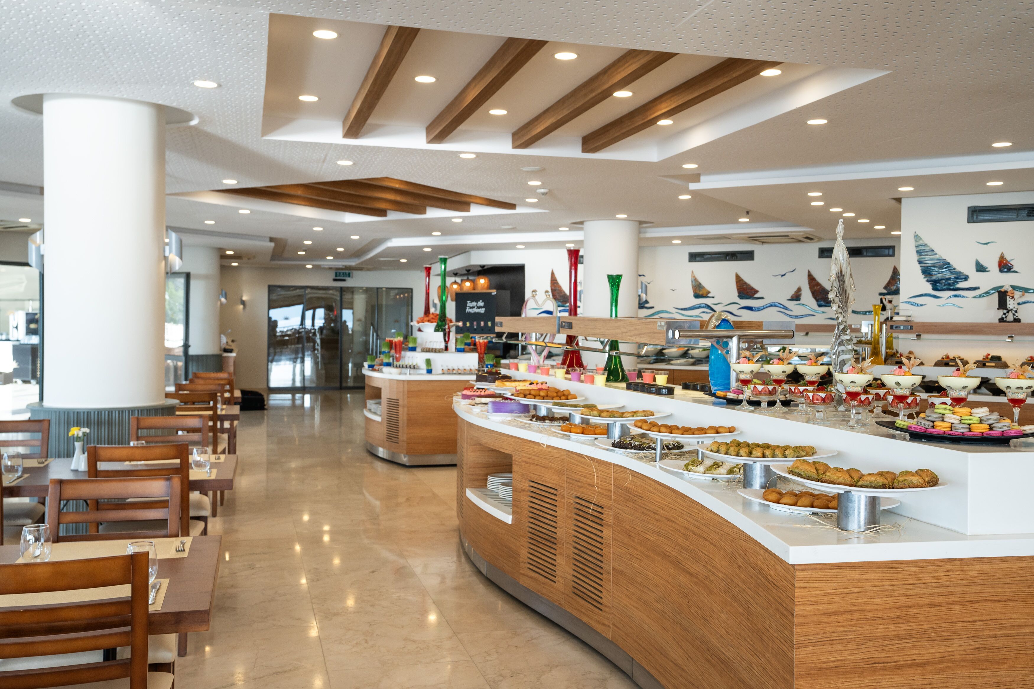 A modern hotel lobby with a buffet area featuring various food options, wooden furniture, and a nautical-themed wall mural.