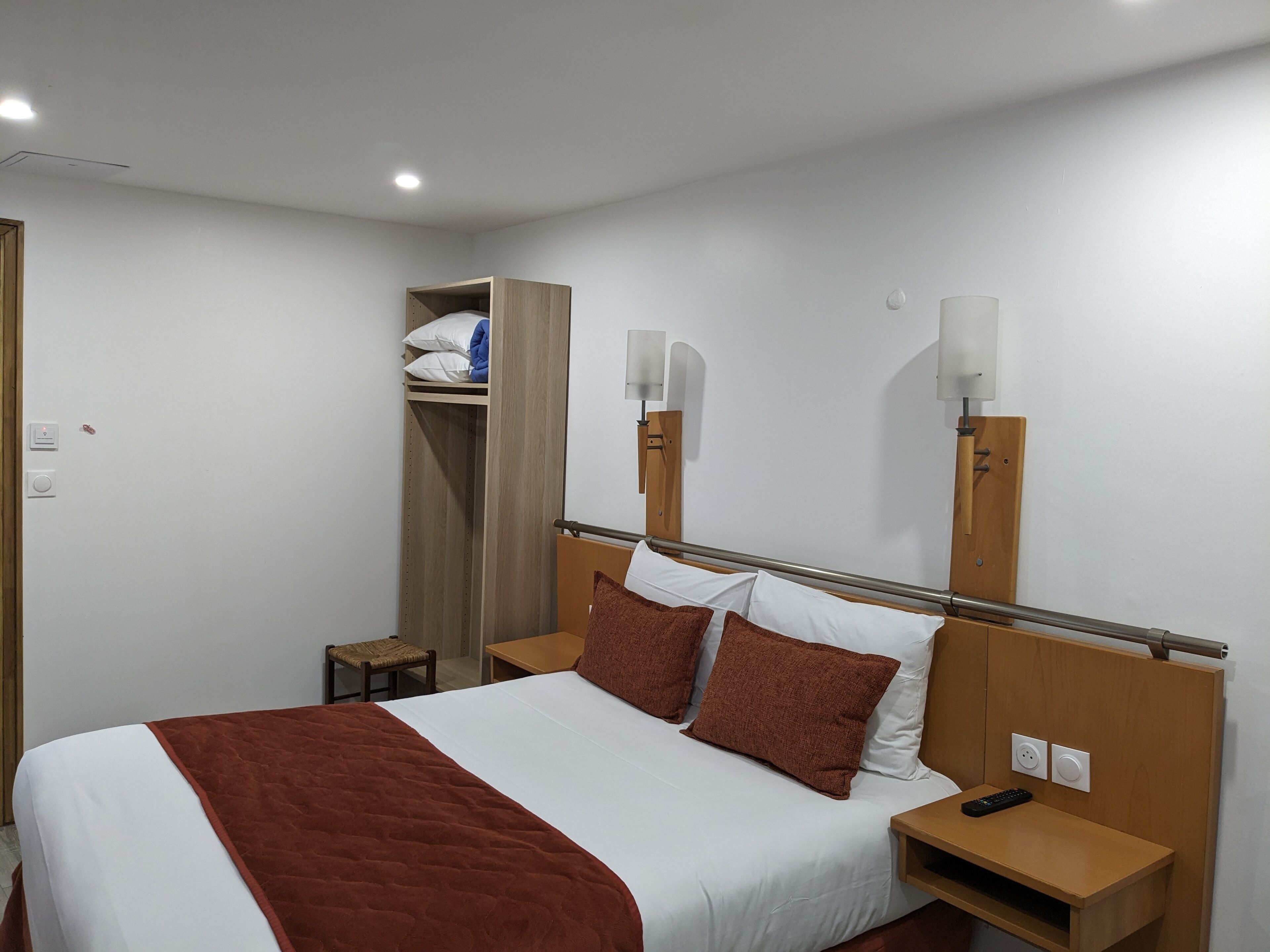 Hotellihuone, jossa on sänky, yöpöytä, yölamppu, vaatekaappi ja pieni jakkara.